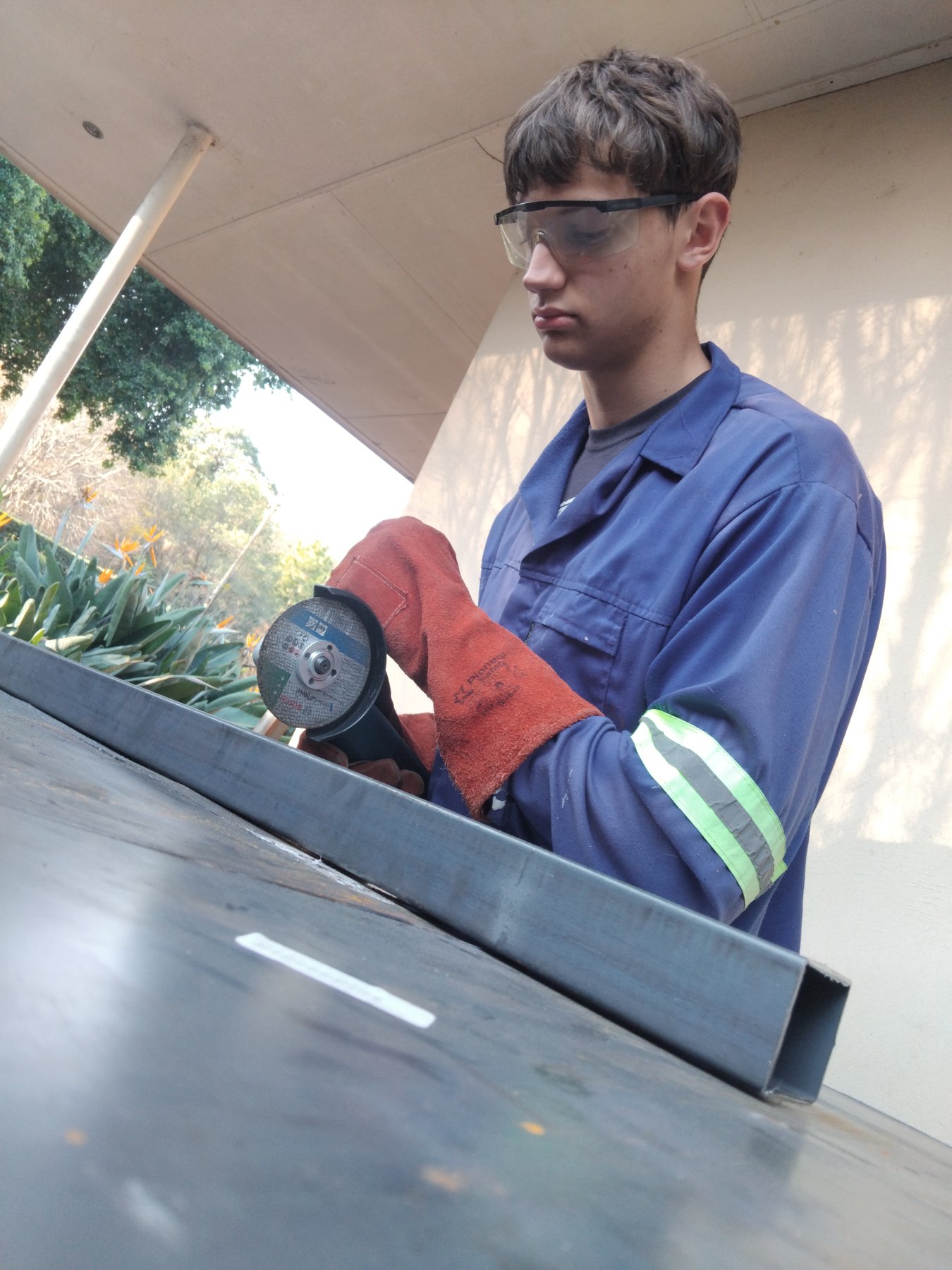 Alex, in overalls and safety glasses, grinding a weld on a steel frame outside the workshop