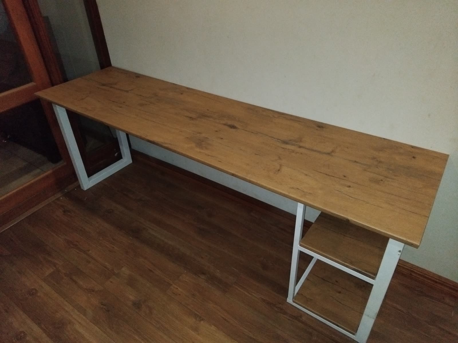 The long oak-look desk against the wall it was built for, white steel frame and a warm oak-look top